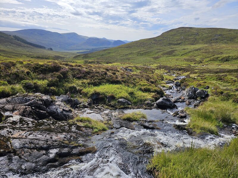 A burn running over dark rock through heather and grass, with a green hill rising behind.