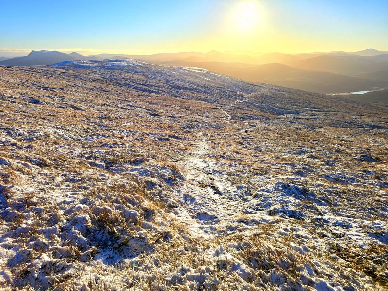 A frosted path running down a broad grassy slope towards a low sun, with hills and a loch in the distance.
