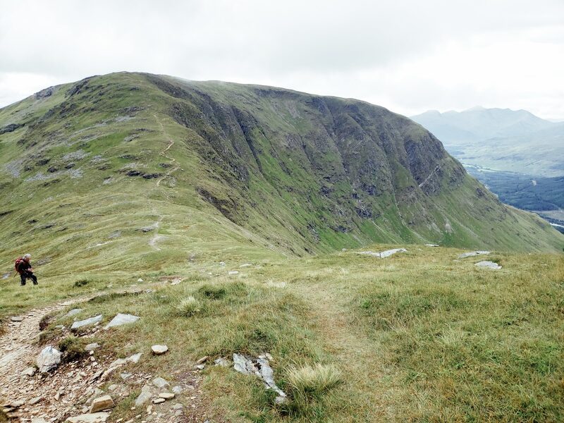 A flat-topped hill with a craggy face seen across a grassy saddle, with a worn path leading down towards it.