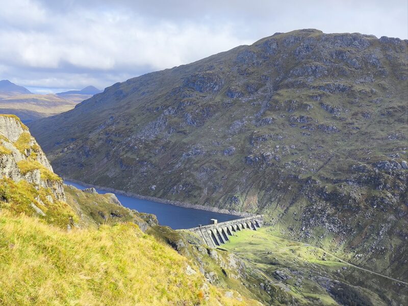 The Loch Sloy dam and its reservoir seen from the grassy slopes of Ben Vane, a steep craggy hillside rising beyond.