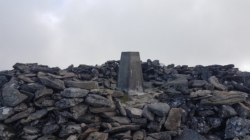 The trig pillar on the summit of Meall Ghaordaidh inside its ring of piled stones, under a pale grey sky.