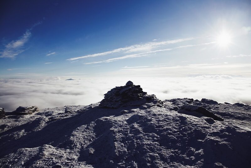 A frosted summit cairn above a sea of cloud, the sun low in a deep blue sky.