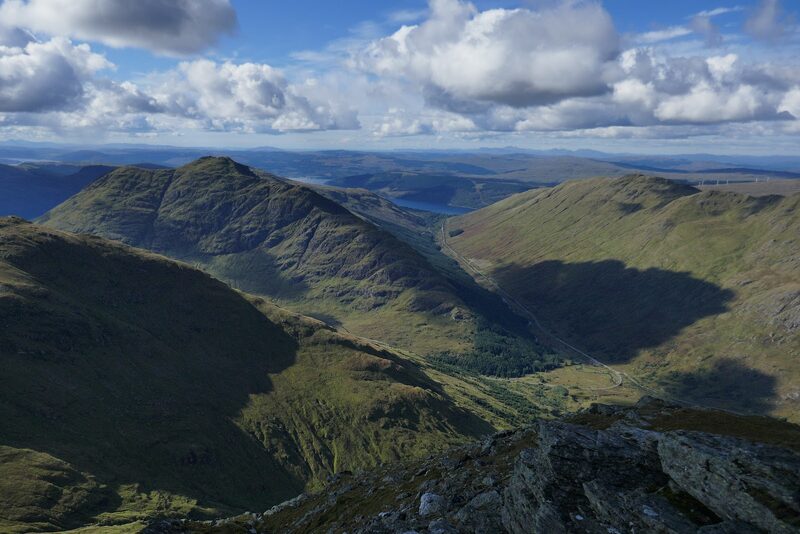 Looking down from the rocks of Beinn Ìme into a deep green glen, a cone-shaped hill across it and water in the distance.