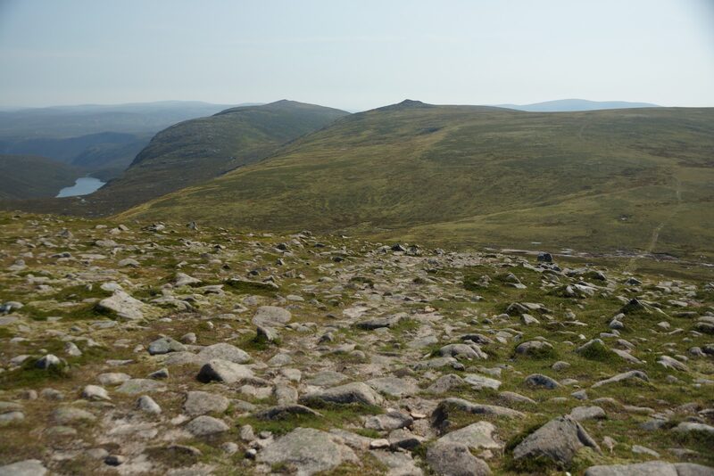 The stony top of Carn an t-Sagairt Mor looking over green slopes to a twin-topped hill, a small loch far below.
