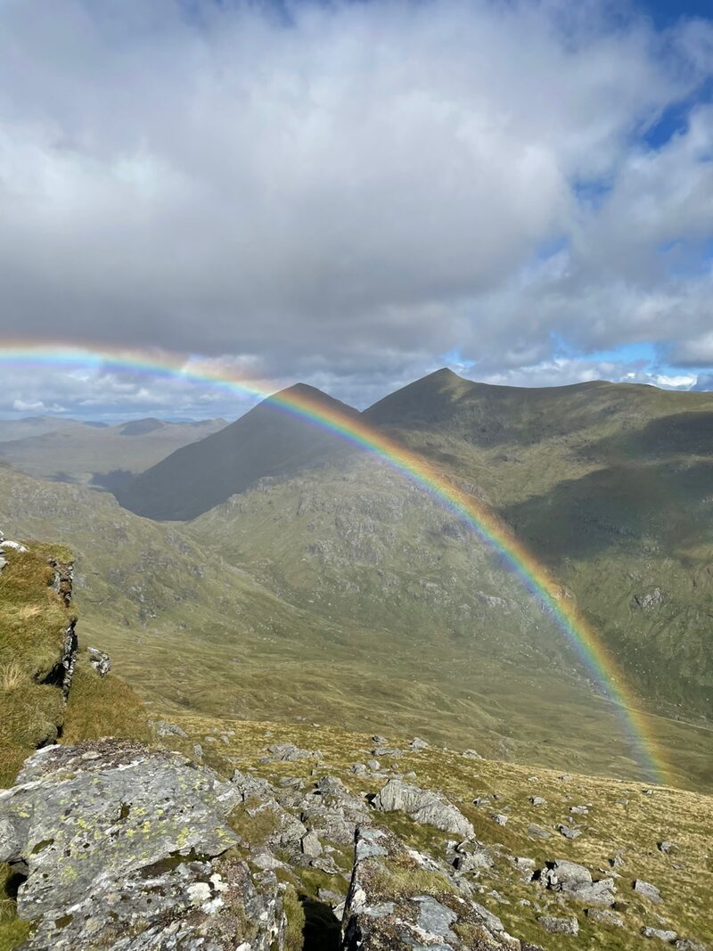 A rainbow arching over a green glen below two pointed hills, seen from rocks high on the hill.