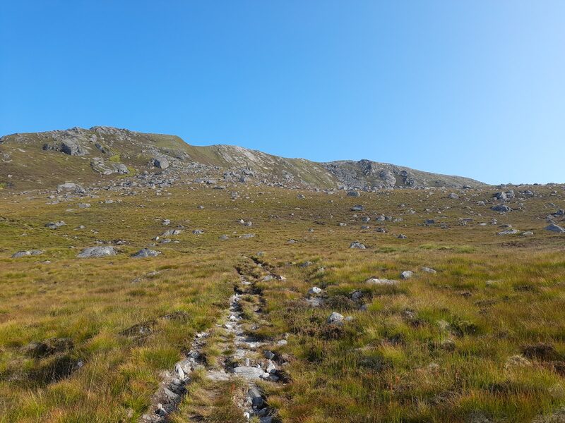 A worn path climbing rough grass towards the rocky top of Càrn Ghluasaid under a blue sky.