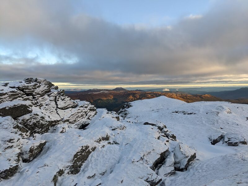 Snow-plastered rocks on a top at first light, brown hills below a band of cloud.