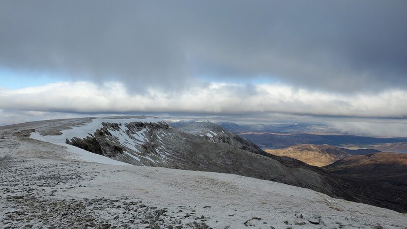 The edge of a high plateau under a dusting of snow, with dark crags falling away below it and sunlit moor beyond.