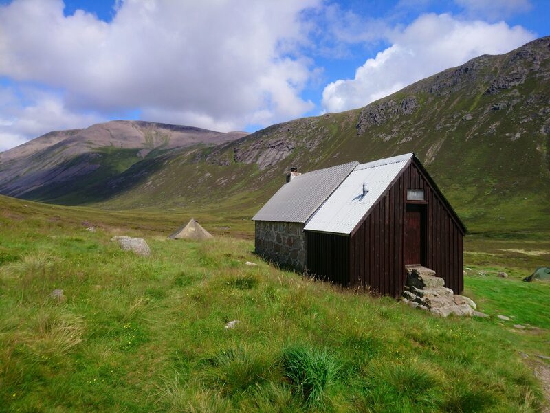 A small stone and timber bothy on the grass, with a steep hillside of crags behind it.