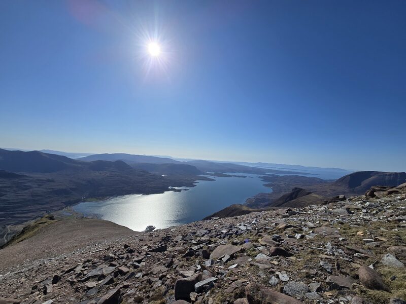 The sea loch and its islands far below a stony slope high on Liathach, into a low bright sun.