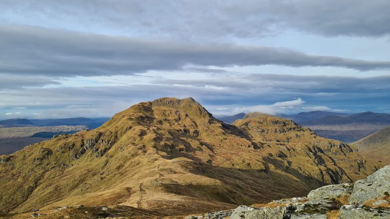 A tawny, craggy hill with a path climbing its ridge to a flattish top, under bands of grey cloud.