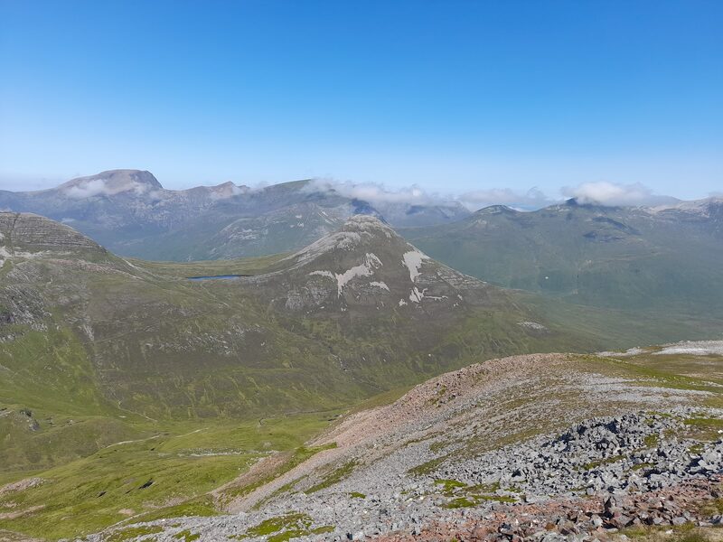 Looking down on the hill's cone and its lochan from a stony shoulder on a clear summer day, with a long high range across the glen behind.