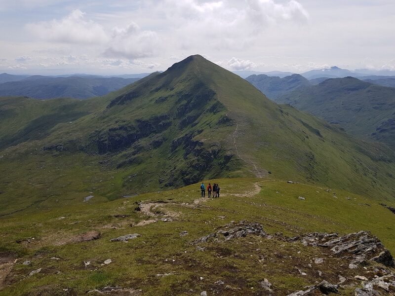 A grassy path dropping to the gap between Ben More and Stob Binnein and climbing the green pyramid beyond, three walkers small in the distance.