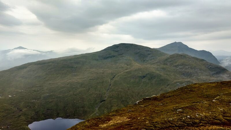 Ben Oss seen whole from the next hill: a broad, rounded top above the dark water of a hill loch, with a sharper peak behind its shoulder under grey cloud.