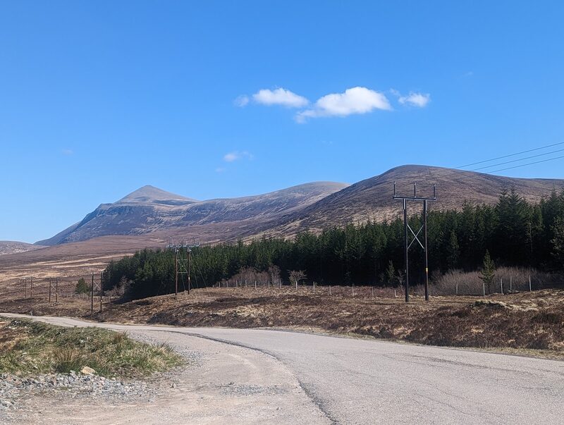 Ben Klibreck's long ridge and pointed top above moor and a block of forestry, seen from a single-track road under a blue sky.