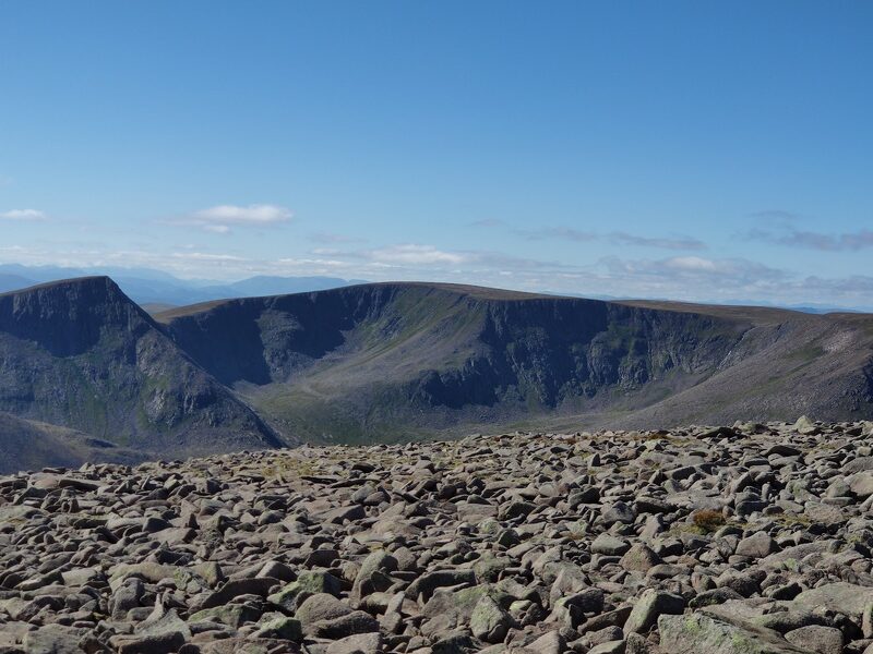 The boulder field of the summit plateau with cliffs and a long corrie wall beyond under a blue sky.