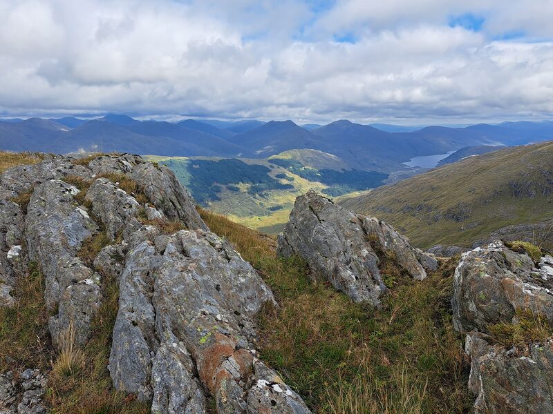 Rocky outcrops high on a ridge, looking down to a forested glen and a distant loch.