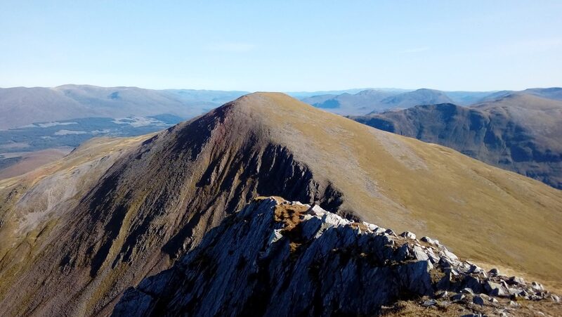 Stob a' Choire Mheadhoin seen across the gap from the rocky top of its twin, dark crags on one flank and grass on the other, a wooded strath and far hills behind