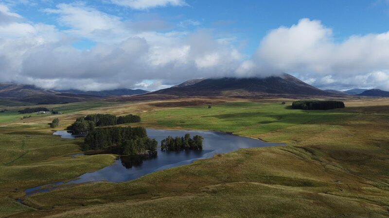 Loch Moraig and its wooded islets from the air, with Càrn Liath dark under cloud beyond the moor.