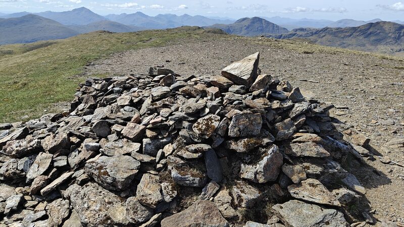 The view from the summit cairn, its slabs filling the foreground, with a row of pointed hills on the skyline beyond the stony top.