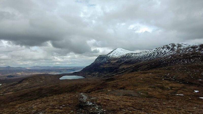 A worn path over brown moor towards Ben Klibreck's upper slopes, dusted with snow, a lochan below and heavy cloud above.