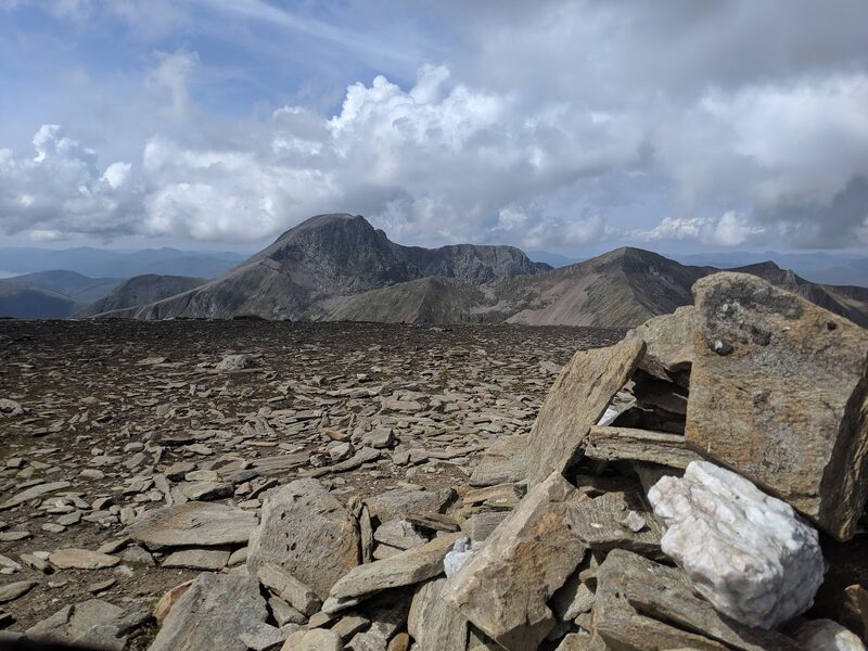 A cairn of slabs with a white quartz block on a flat top of slaty stones, and a dark, steep-faced mountain beyond under piled cloud.