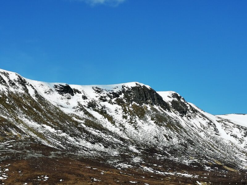 A line of dark crags under patchy snow along the rim of a hill, beneath a clear blue sky.