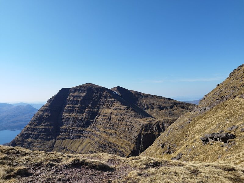 A sheer, tiered sandstone top of Beinn Alligin in shadow, seen across a gap from higher on the mountain, with the loch far below.