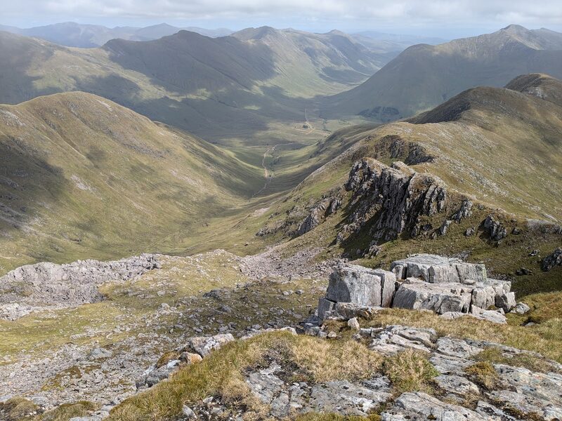 Pale rock slabs and a crag on a grassy ridge, with a long green glen and a thin road far below.