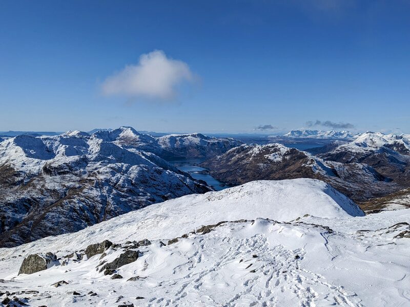 A winter view from a snow-covered ridge down to a loch between white, craggy mountains under a blue sky.