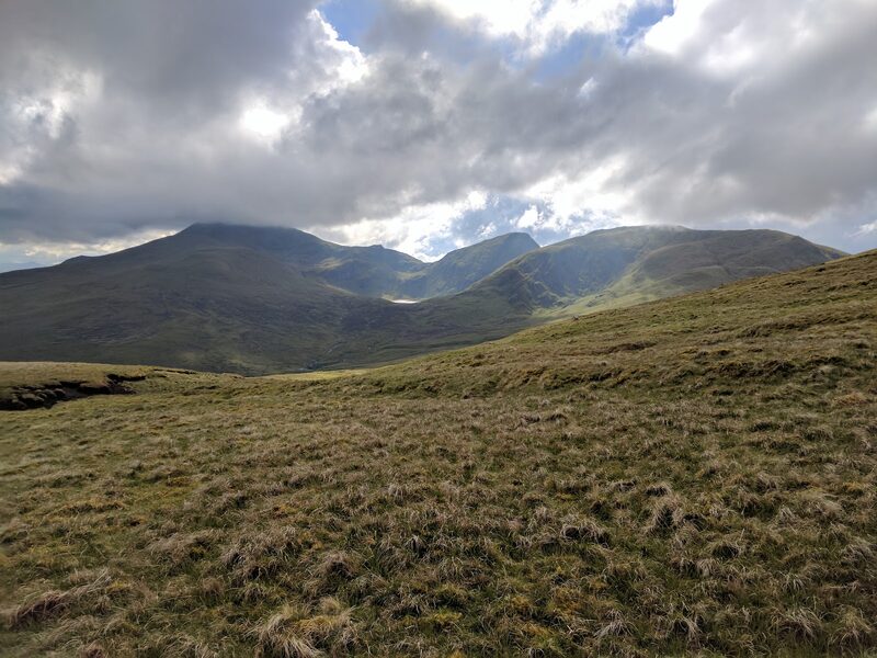 A slope of rough grass with three higher hills beyond, their tops under heavy cloud.
