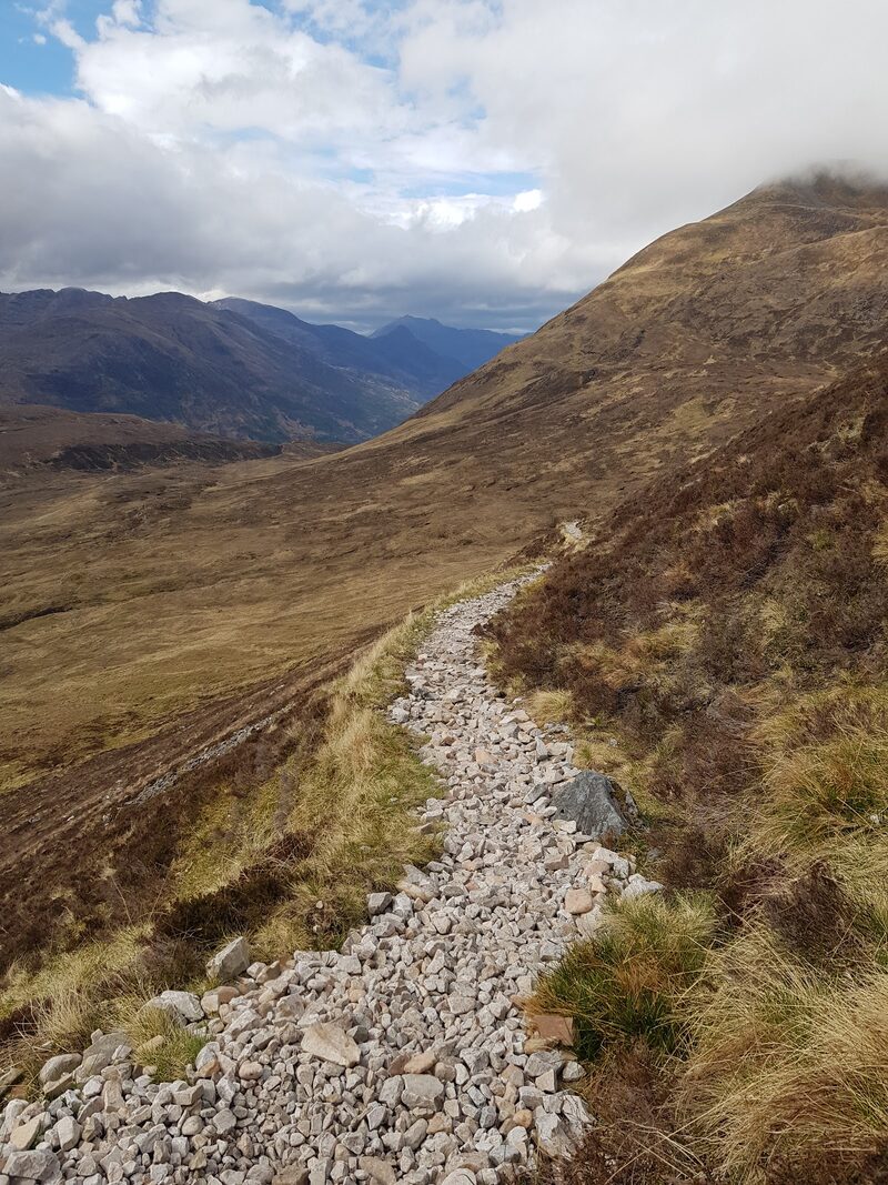 A path of pale stones crossing a heather slope, the hillside rising into cloud on the right and a wide glen falling away on the left.