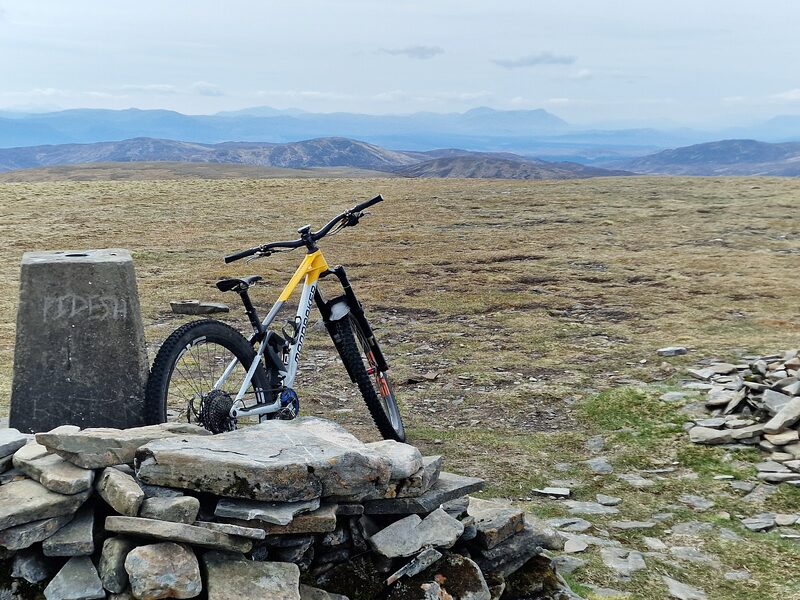 The trig pillar and its low stone shelter on the wide grassy top, a mountain bike leaning on the stones, blue hills far off.
