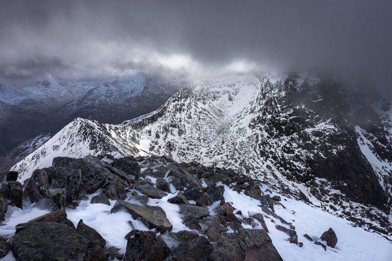 Snow-covered boulders on Càrn Mòr Dearg and the narrow ridge beyond, under dark cloud with light on the far slopes.
