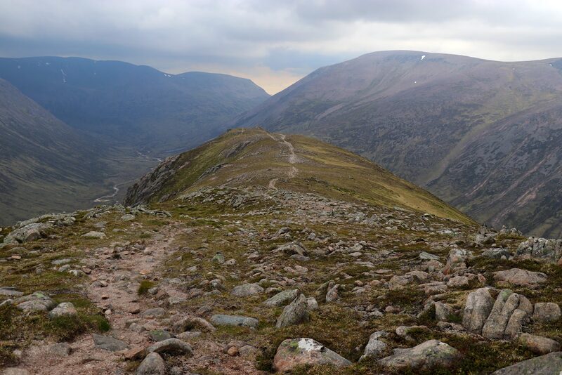 A stony path running out along the crest of a narrow ridge, with a deep glen falling away on either side under heavy cloud.