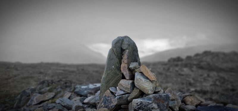 A summit cairn on Druim Shionnach topped by a tall upright stone, with hills dim in the mist behind.
