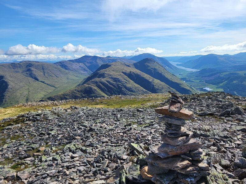 A small cairn of flat stones on the stony top, the ground falling away beyond it to a green glen floor and a sea loch, hills on either side under a blue sky.