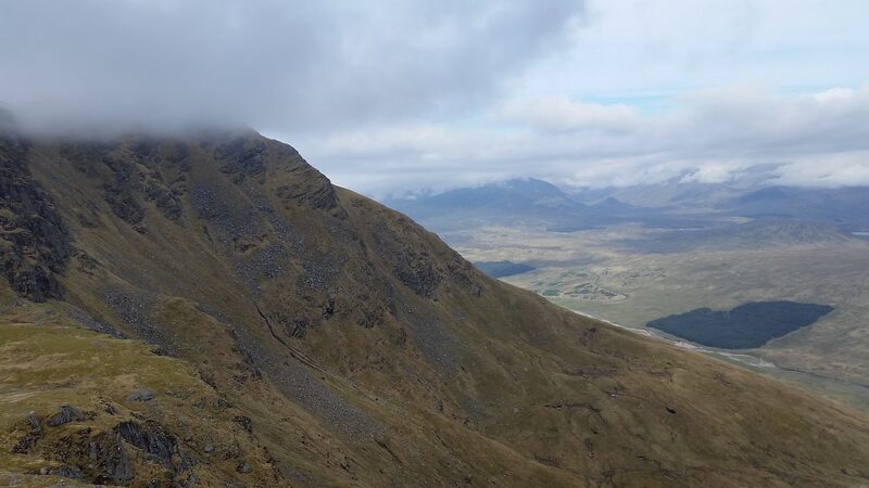 A dark, craggy hillside with cloud on its top, falling away to a wide moor, a plantation and far hills.