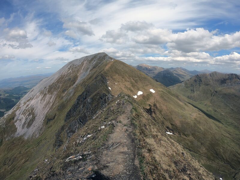 The top of the hill with pale screes down its left flank and small snow patches, seen along a grassy crest with a worn path.
