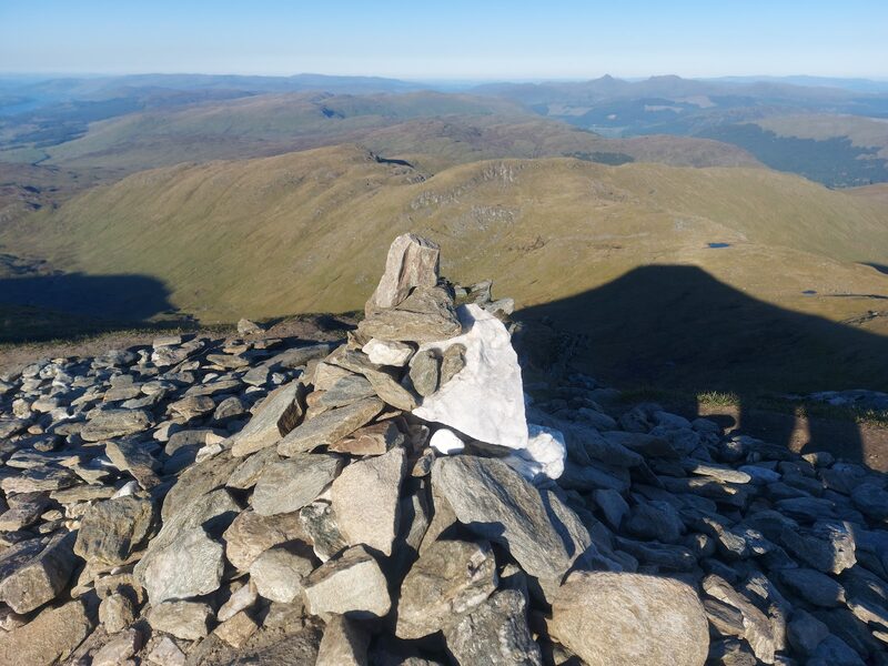 A summit cairn of grey stones topped with white quartz, the hill's own shadow thrown across the glen below.