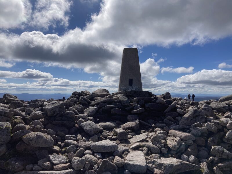The trig pillar on its built cairn of boulders under big cloud, with two figures far off.
