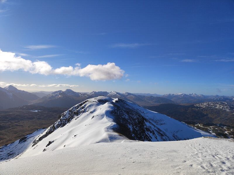 Ben Challum's summit ridge under snow, dipping to a lower top with a blue sky and far ranges beyond.