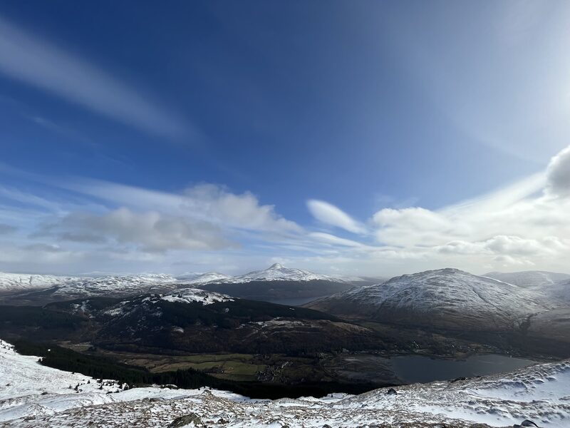 Snow on the slopes of Beinn Narnain, the head of the sea loch far below and white hills beyond under a blue sky.
