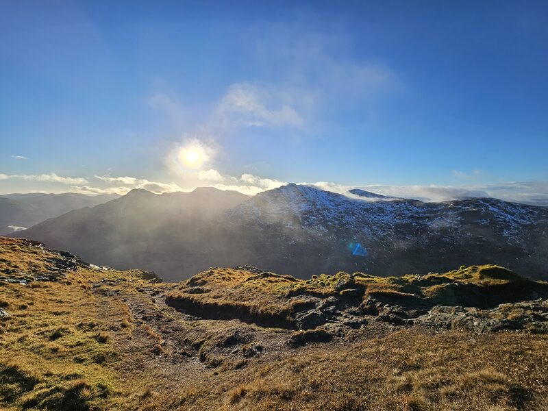Tussocky high ground on Ben Vorlich against a low sun, with snow-dusted hills across the glen.