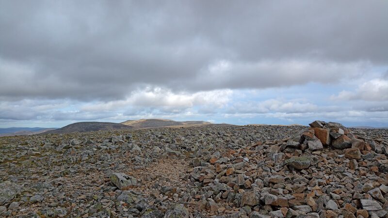 The wide top of Càrn an Tuirc, a field of broken stones with a low cairn of reddish rock, far rounded hills under grey cloud.