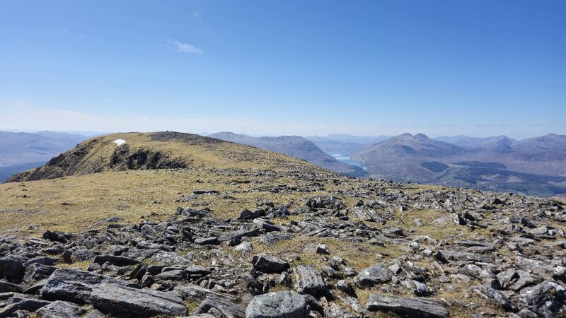 The stony summit plateau in clear sun, the ridge running on to a second top with a craggy flank and a patch of old snow, and a loch between hills beyond.