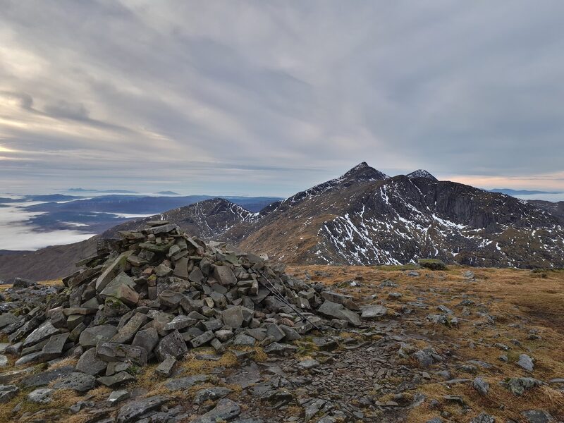 Ben Cruachan's pointed summit and its neighbouring top, streaked with old snow, seen along the ridge from a stone cairn, low cloud over the glens beyond.