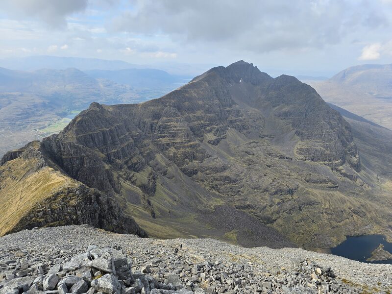 From grey summit scree, a dark corrie with a lochan and Liathach's other Munro beyond it, the sea loch far below to the left.