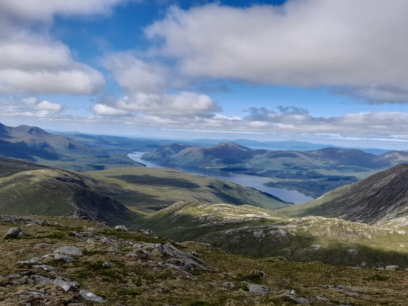 The view from high on the hill over green, slabby knolls to a long loch winding between low hills, the sea and islands beyond under broken cloud.
