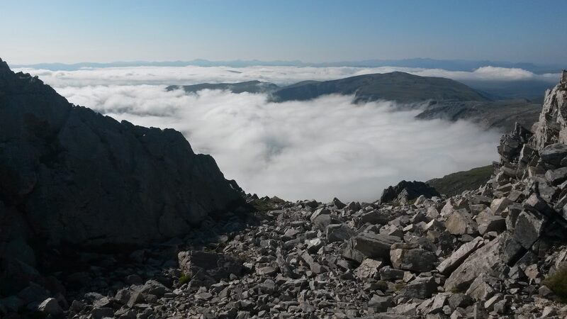 A slope of shattered rock high on Ben More Assynt, looking out over a sea of cloud with hilltops standing clear of it.